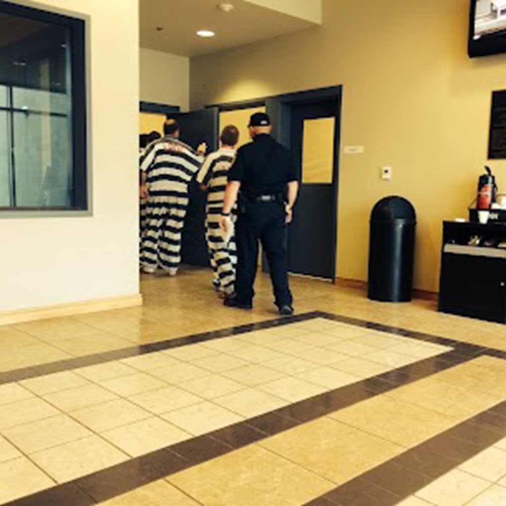 Prisoners in black and white striped jumpsuits exiting jailhouse followed by guard.