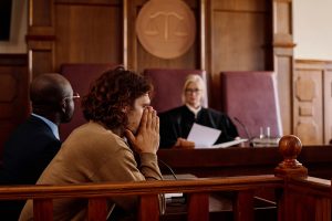 Young anxious male suspect covering his face by hands while sitting in front of camera against judge passing judgment during trial session.