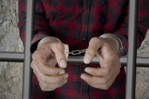 imgmaleprisonerwithhandcuffsjail Affordable Bail Affordable Bail Person in a red plaid shirt with hands in handcuffs gripping a cell bar
